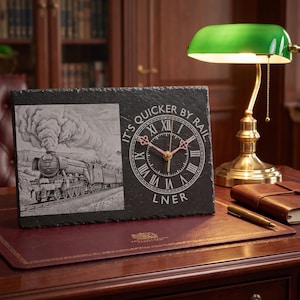 May include: A rectangular black slate clock featuring an engraved steam locomotive on the left and a circular clock face on the right. The text IT'S QUICKER BY RAIL and LNER is etched around the clock face, which has Roman numerals and gold-coloured hands.