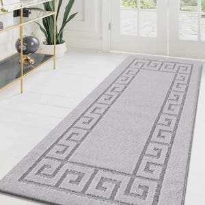 May include: A rectangular light grey bath mat featuring a Greek key border design. The mat is made of a soft, plush material, suitable for bathrooms or entryways. The mat measures approximately 152 cm in length.