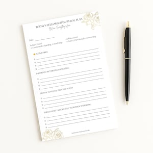 May include: A white notepad with gold floral accents and the text "TODAY'S FELLOWSHIP SURVIVAL PLAN". A black pen with gold accents rests beside the notepad. The notepad includes sections for mood, caffeine level, and notes.