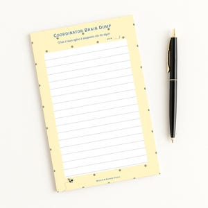 May include: A pale yellow notepad with the text "COORDINATOR BRAIN DUMP" and lined pages. A black pen with gold accents rests beside the notepad. The notepad has a dotted border and the text "Write it down before it disappears into the abyss".