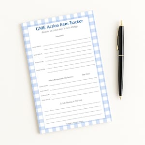 May include: A notepad with a blue and white gingham border and the text "GME Action Item Tracker." The notepad has lines for action items, a section for who is responsible, and a due date. A black pen with gold accents rests beside the notepad.