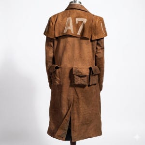 Puede incluir: Abrigo largo de ante marrón con múltiples bolsillos y una abertura en la espalda. El abrigo presenta un detalle tipo capa en los hombros y el número "A7" en blanco en la parte superior de la espalda. El abrigo se exhibe en un maniquí.