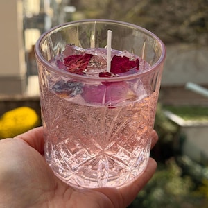 Può includere: Una candela rosa in un bicchiere trasparente a motivi. La candela contiene un liquido rosa e petali di rosa. Uno stoppino bianco è centrato. Il bicchiere è tenuto in mano. La candela è alta circa 8 cm.