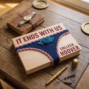 May include: A hardcover book titled "IT ENDS WITH US" by Colleen Hoover, wrapped with a blue crocheted design with pink trim and a teal flower. The book rests on a wooden table with a vintage watch and a gold pen.