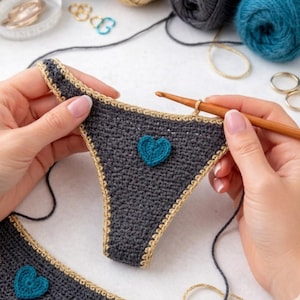 May include: Hand-crocheted dark grey underwear with a teal heart motif. The edges are trimmed with a light brown yarn. A wooden crochet hook is being used. The image shows the item being crafted, with yarn and other crafting supplies in the background.