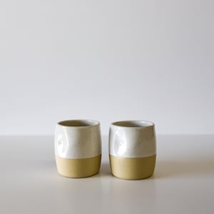 May include: Two ceramic tumblers with a speckled white upper half and a solid yellow-beige lower half. The cups have a smooth, rounded shape and are set against a plain white background.