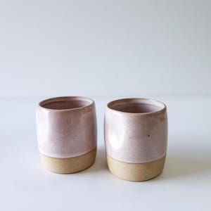 May include: Two handmade ceramic cups. The cups are a soft pink color on the top half and a natural tan color on the bottom half. The cups have a smooth, rounded shape and are likely used for drinking.