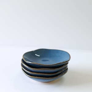 May include: A stack of four small, round ceramic dishes with a smooth, matte blue glaze on the interior and a dark, contrasting rim. Each dish features an organic, slightly irregular edge, creating a layered appearance in this set of stoneware.
