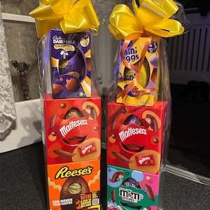 Pode incluir: Duas cestas de presente de P&aacute;scoa embrulhadas em celofane transparente, cada uma encimada por um grande la&ccedil;o amarelo. Uma cesta inclui um ovo Cadbury Dairy Milk Caramel, Maltesers e um ovo Reese's. A outra tem Mini Eggs, Maltesers e M&M's minis.