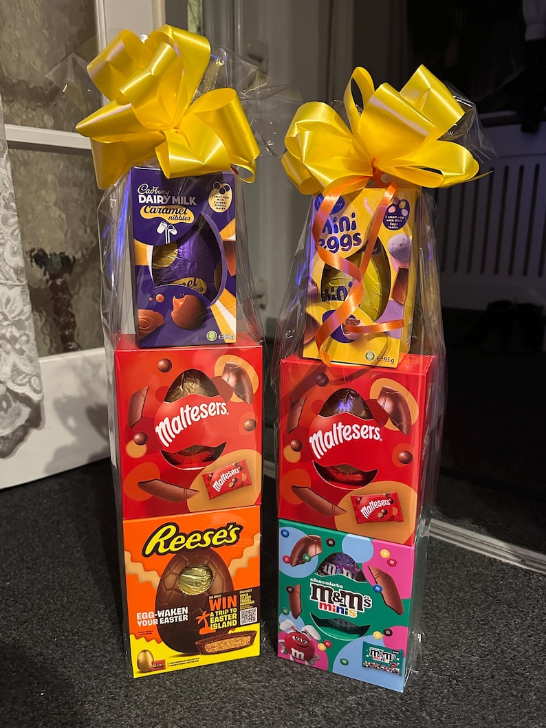 Pode incluir: Duas cestas de presente de P&aacute;scoa embrulhadas em celofane transparente, cada uma encimada por um grande la&ccedil;o amarelo. Uma cesta inclui um ovo Cadbury Dairy Milk Caramel, Maltesers e um ovo Reese's. A outra tem Mini Eggs, Maltesers e M&M's minis.