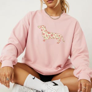 Könnte beinhalten: Ein hellrosa Langarm-Sweatshirt mit Rundhalsausschnitt, das auf der Vorderseite eine zentrale Grafik der Silhouette eines Dackels zeigt, die mit einem cremefarbenen Blumenmuster aus kleinen roten Rosen und grünen Blättern gefüllt ist.