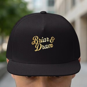 May include: Black flat-peak cap featuring the gold text "Briar & Dram" in a script font. The cap has a structured crown and a flat peak. A classic style of hat, suitable for casual wear.
