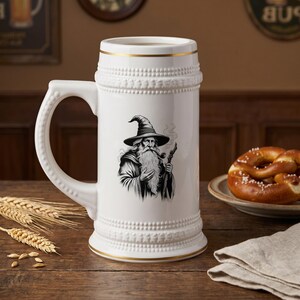 May include: White ceramic beer stein with a handle, featuring a black and white illustration of a wizard with a long beard and hat. The stein has gold trim and decorative detailing. A pretzel sits on a plate nearby.
