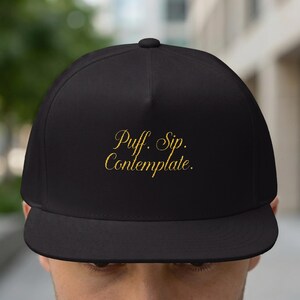 May include: Black flat-brimmed baseball cap with the embroidered text "Puff. Sip. Contemplate." in gold script. The cap has a structured fit and a curved brim, suitable for casual wear.