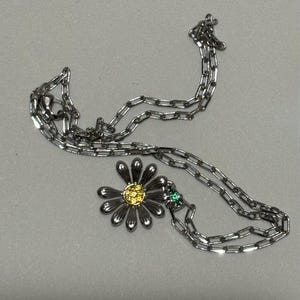 Puede incluir: Collar con colgante de margarita plateada con centro amarillo y piedra verde. La cadena es de estilo eslabón delicado. El collar se muestra sobre una superficie gris claro.