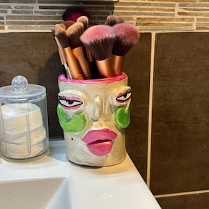 Könnte beinhalten: Ein zylindrischer Make-up-Pinselhalter in Gesichtsform mit cremefarbener Basis, leuchtend pinken Lippen, grünen Wangenakzenten und dunklen Augen. Der Halter ist mit verschiedenen Make-up-Pinseln mit braunen Borsten und kupferfarbenen Griffen gefüllt.