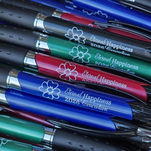 May include: A close-up of several pens in various colors, including black, green, red, and blue. Each pen has a silver tip and a barrel with the text "Eternal Happiness 2026 Convention" and a floral design.