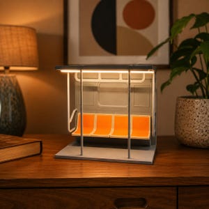 May include: A miniature subway car interior lamp featuring four bright orange moulded plastic seats, a grey frame, and a white overhead grab rail. The structure is illuminated by a warm LED light strip integrated into the top ceiling panel.