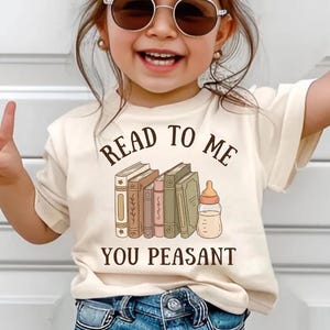 Op de afbeelding: Een crèmekleurig T-shirt met de tekst "READ TO ME YOU PEASANT" in het zwart. Het shirt heeft een afbeelding van verschillende boeken en een babyfles. De persoon op de foto draagt een zonnebril en een blauwe spijkerbroek.