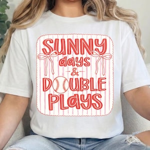 May include: White t-shirt with a red and pink graphic. The graphic reads "SUNNY days & DOUBLE PLAYS" with a baseball illustration. The design includes bows and vertical stripes, creating a playful, sporty aesthetic.
