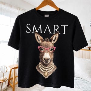 May include: Black t-shirt featuring a donkey wearing pink glasses and a pearl necklace. The word "SMART" is printed in white above the donkey's head. The shirt is hanging on a wooden hanger.