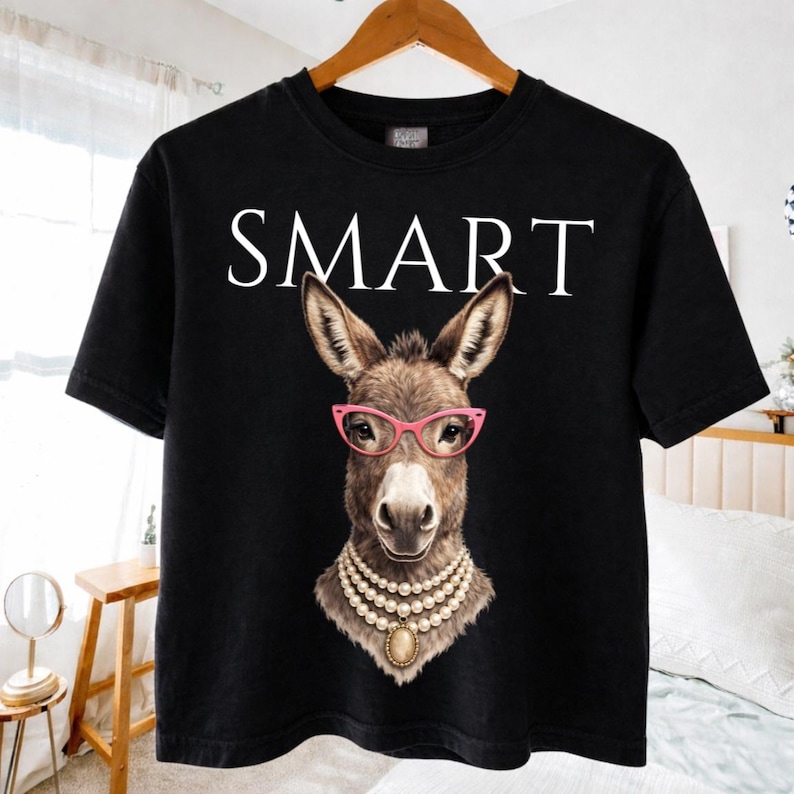 May include: Black t-shirt featuring a donkey wearing pink glasses and a pearl necklace. The word "SMART" is printed in white above the donkey's head. The shirt is hanging on a wooden hanger.