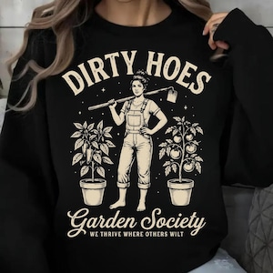 Op de afbeelding: Zwarte sweatshirt met een afbeelding van een vrouw met een schoffel, geflankeerd door potplanten. De tekst luidt "DIRTY HOES" erboven en "Garden Society" eronder, met de slogan "WE THRIVE WHERE OTHERS WILT".