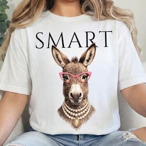 Puede incluir: Camiseta blanca con la palabra "SMART" en letras negras. La camiseta presenta una ilustración detallada de un burro con gafas rosas y un collar de perlas. El burro tiene pelaje marrón.