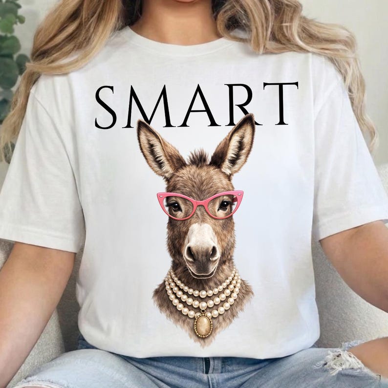 May include: White t-shirt with the word "SMART" in black block letters. The shirt features a detailed illustration of a donkey wearing pink glasses and a pearl necklace. The donkey has brown fur.
