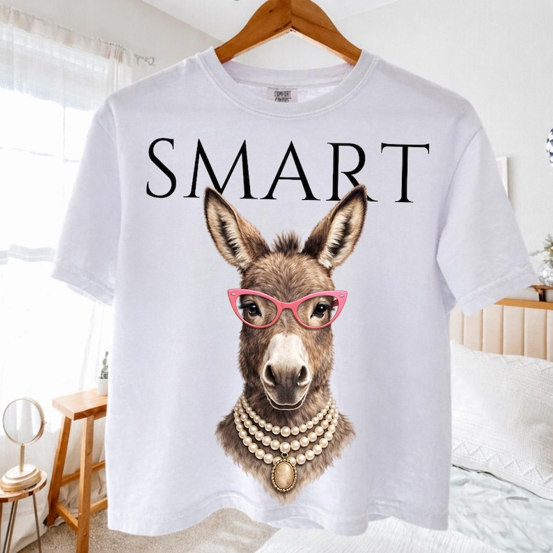 May include: White t-shirt with the word "SMART" in black block letters. The shirt features a detailed illustration of a donkey wearing pink glasses and a pearl necklace. The donkey has brown fur and is centered on the shirt.