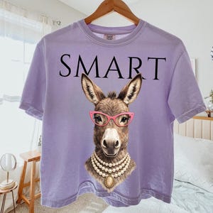 May include: A lilac-colored t-shirt with the word "SMART" printed across the top. The shirt features a graphic of a donkey wearing pink glasses and a pearl necklace. The donkey graphic is centered on the shirt.