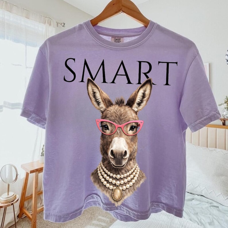 May include: A lilac-colored t-shirt with the word "SMART" printed across the top. The shirt features a graphic of a donkey wearing pink glasses and a pearl necklace. The donkey graphic is centered on the shirt.