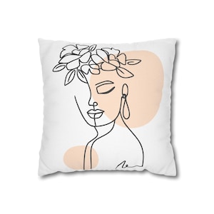 May include: White square throw pillow featuring a minimalist line drawing of a woman's face with a floral crown and teardrop earrings. The design includes a peach-colored background element. The pillow is a decorative home accent.