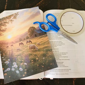 Könnte beinhalten: Ein rechteckiger Druck mit einer ländlichen Landschaft, in der Schafe bei Sonnenuntergang auf einer Blumenwiese grasen, platziert auf einem offenen Buch neben einer Schere mit blauem Griff und einer Rolle weißem Klebeband.