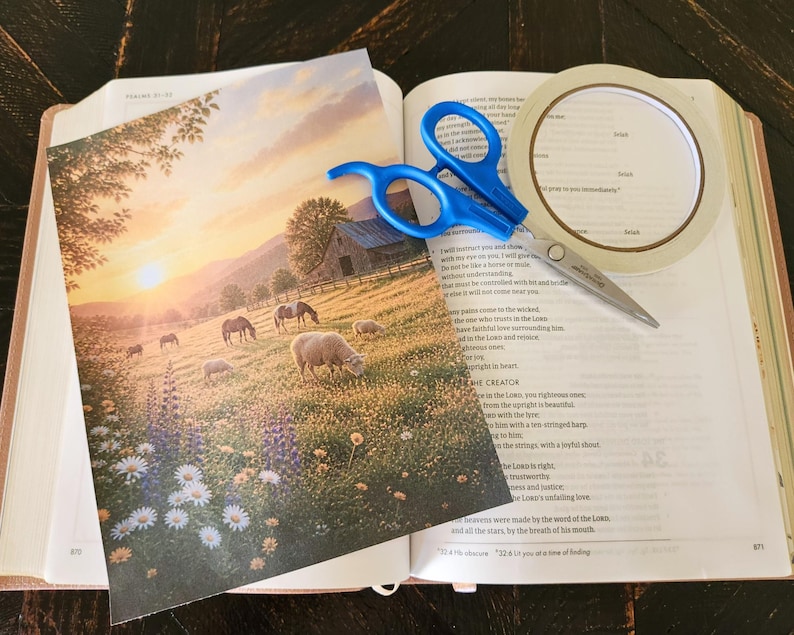 Peut inclure: Une image imprim&eacute;e repr&eacute;sentant un paysage pastoral avec des moutons broutant dans un champ de fleurs sauvages au coucher du soleil, pos&eacute;e sur un livre ouvert avec une paire de ciseaux &agrave; poign&eacute;es bleues et un rouleau de ruban adh&eacute;sif blanc.