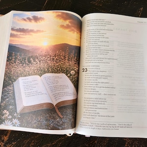 Peut inclure: Une Bible ouverte affichant le Psaume 23, avec une illustration pleine page en couleur d'un coucher de soleil sur un champ herbeux parsem&eacute; de fleurs sauvages. Le texte inclut le titre Le Bon Berger et les versets du Psaume 23 en noir sur papier blanc.