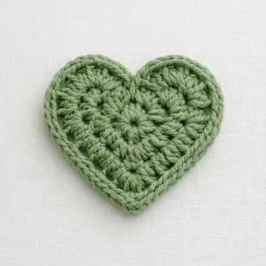May include: A sage green crochet heart, handmade with a textured stitch and a defined border. The craft item is displayed on a white background. Ideal for Valentine's Day or other craft projects. The heart is approximately 10 cm wide.
