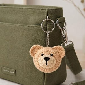 Op de afbeelding: Olijfgroene crossbodytas met een gehaakte teddybeer-bedel. De bedel is met een zilveren ketting en ring aan de tas bevestigd. De tas heeft een voorvak en de tekst "THE BEAR CHARM".