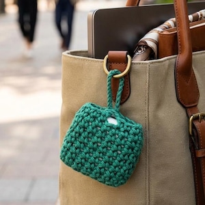 May include: A green, crocheted square case with a loop, hanging from a tan tote bag with brown leather handles. The case appears to hold a pair of white earbuds. The bag contains a laptop and other items.