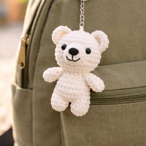 May include: A small, white crocheted teddy bear keychain with black eyes and a black nose hangs from a silver chain attached to a green backpack. The bear has a smiling face and is made of a soft, textured yarn.