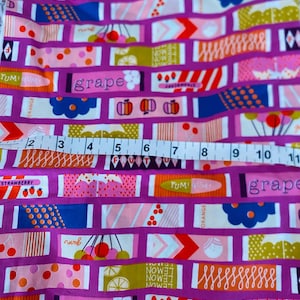 May include: A close-up of a pink and purple fabric with a repeating pattern of colorful fruit and the word "grape". The fabric is measured with a tape measure.