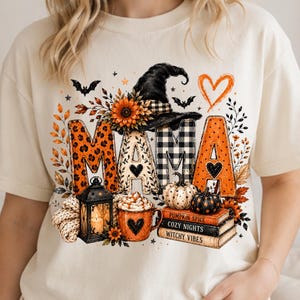 Può includere: Una t-shirt color crema con una grafica che riporta la parola MAMA in vari motivi, un cappello da strega, un girasole, pipistrelli, un cuore, una lanterna, una tazza di caffè, zucche e libri con la scritta PUMPKIN SPICE, COZY NIGHTS, WITCHY VIBES.