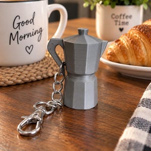 May include: A gray, 3D-printed moka pot keychain featuring a classic octagonal base, a handle, and a hinged lid. The keychain includes a silver-toned metal split ring and a lobster claw clasp attached to the handle of the miniature coffee maker.