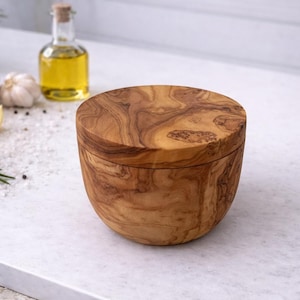 Pode incluir: Um saleiro de madeira com tampa, mostrando um padrão de grão de madeira natural. O recipiente redondo é feito de madeira castanha clara. A imagem também inclui ingredientes como limões, alecrim e alho, sugerindo um tema culinário.