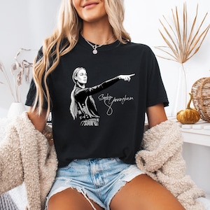 May include: A black short-sleeved t-shirt featuring a white graphic illustration of a person with a ponytail pointing to the right. The text "Sophie Cunningham" is printed in a white cursive script below the illustration on the front of the shirt.