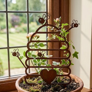 May include: A rusty brown metal beehive-shaped plant trellis with bee and heart cutouts. The trellis is in a terracotta pot with soil and greenery, near a window. The design is ideal for climbing plants.