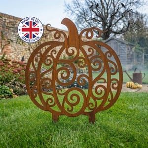May include: A rust-coloured metal garden stake shaped like a pumpkin with intricate swirling cutouts. The pumpkin features a prominent stem and a decorative openwork design throughout its body, standing upright on two prongs in the grass.