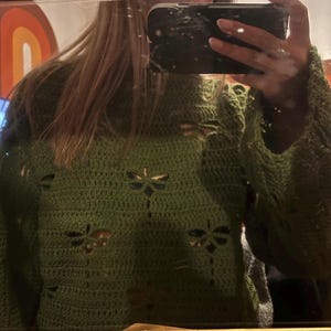 Può includere: Un maglione verde all'uncinetto con ritagli a forma di libellula. Il maglione ha maniche lunghe e un collo alto. Il punto all'uncinetto crea un aspetto strutturato. I ritagli di libellula aggiungono un elemento decorativo al capo.