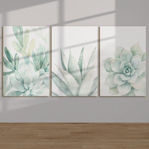 Pode incluir: Um tríptico de pinturas em aquarela com plantas suculentas em tons de verde e azul. A obra de arte é exibida sobre um fundo branco, com os detalhes das plantas claramente visíveis. As pinturas são montadas em uma parede acima de um piso de madeira.