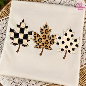 May include: A cream-coloured t-shirt featuring three maple leaf graphics arranged horizontally. The first leaf has a black and cream chequered pattern, the second has a brown and black leopard print, and the third has black polka dots on cream.